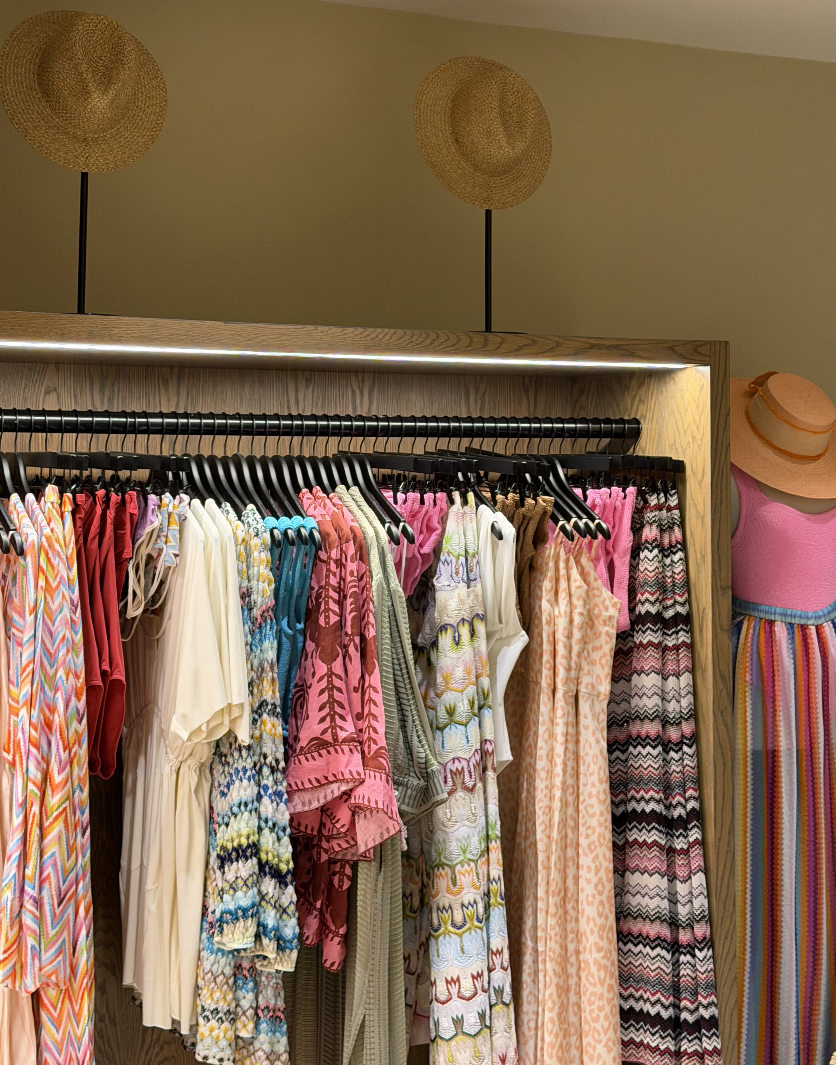 Colorful premium resort apparel arranged on an illuminated boutique rack with sculptural straw hats