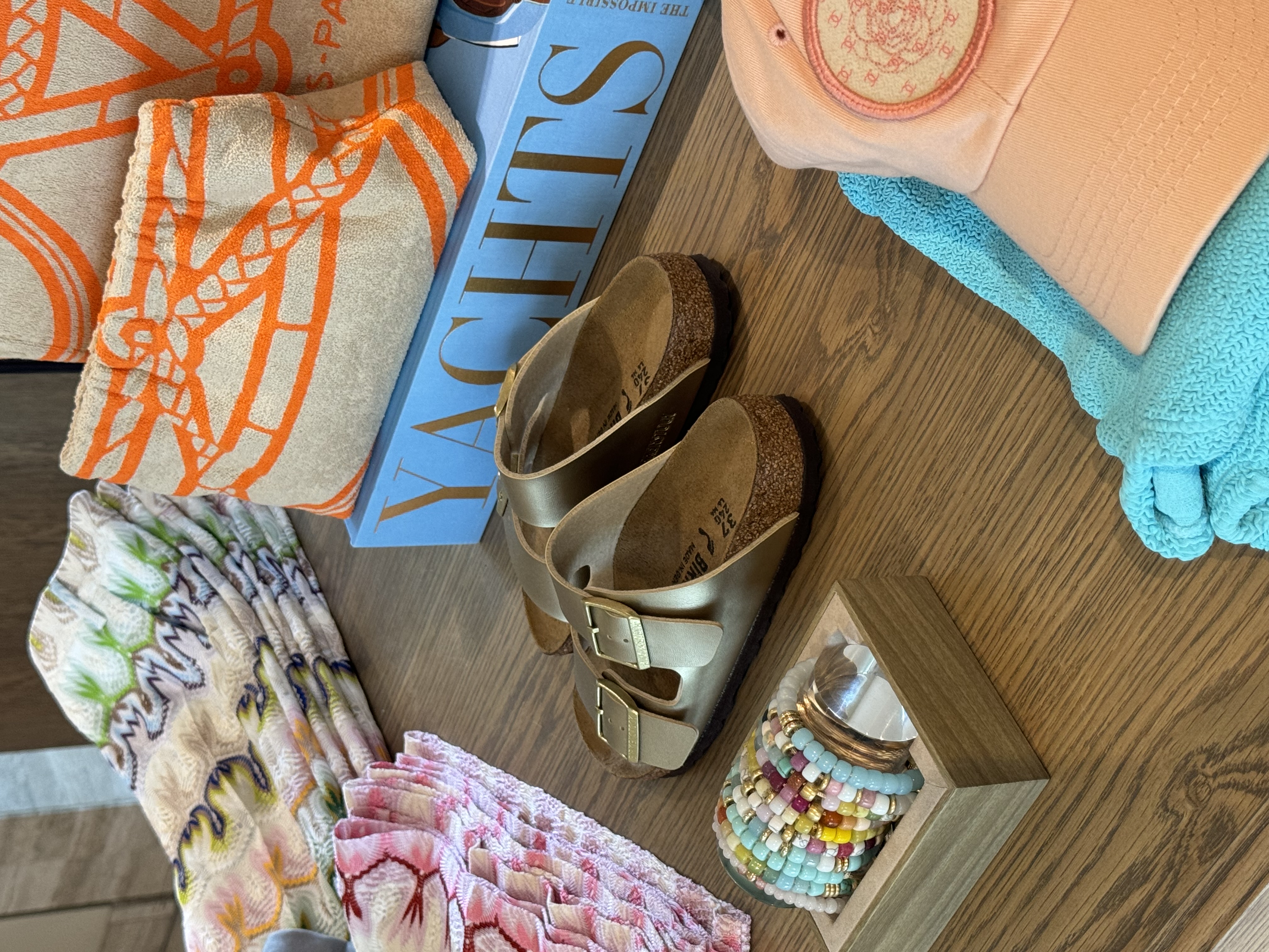 Resort accessories, patterned bags, sandals, bracelets, and folded apparel styled on a warm wood table