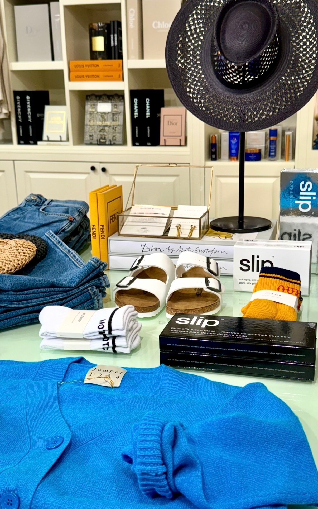 Blue knitwear, denim, books, beauty products, footwear, and a black sculptural hat styled on a boutique table